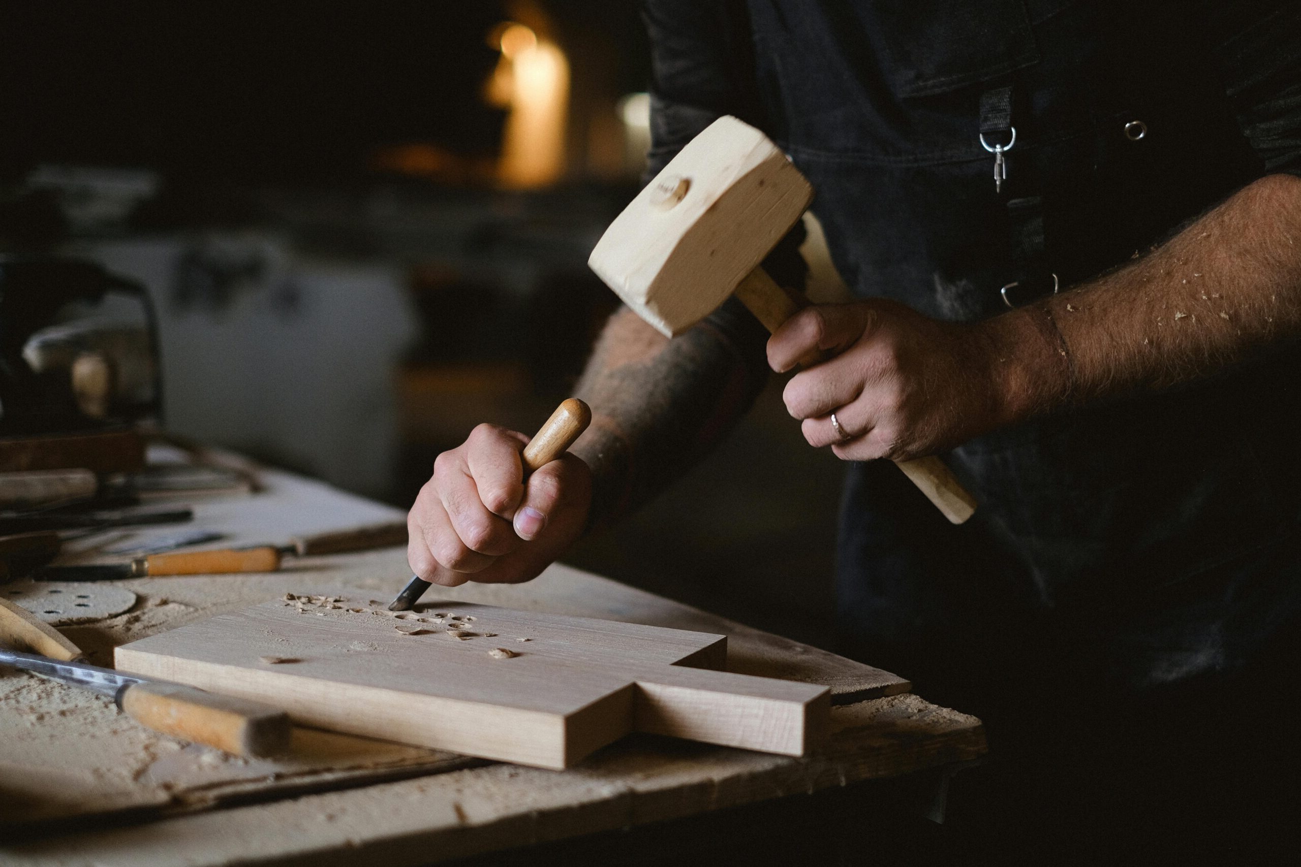 Unrecognizable male artisan using wooden hammer and chisel to carve lumber plank while working at table with special instruments on blurred background