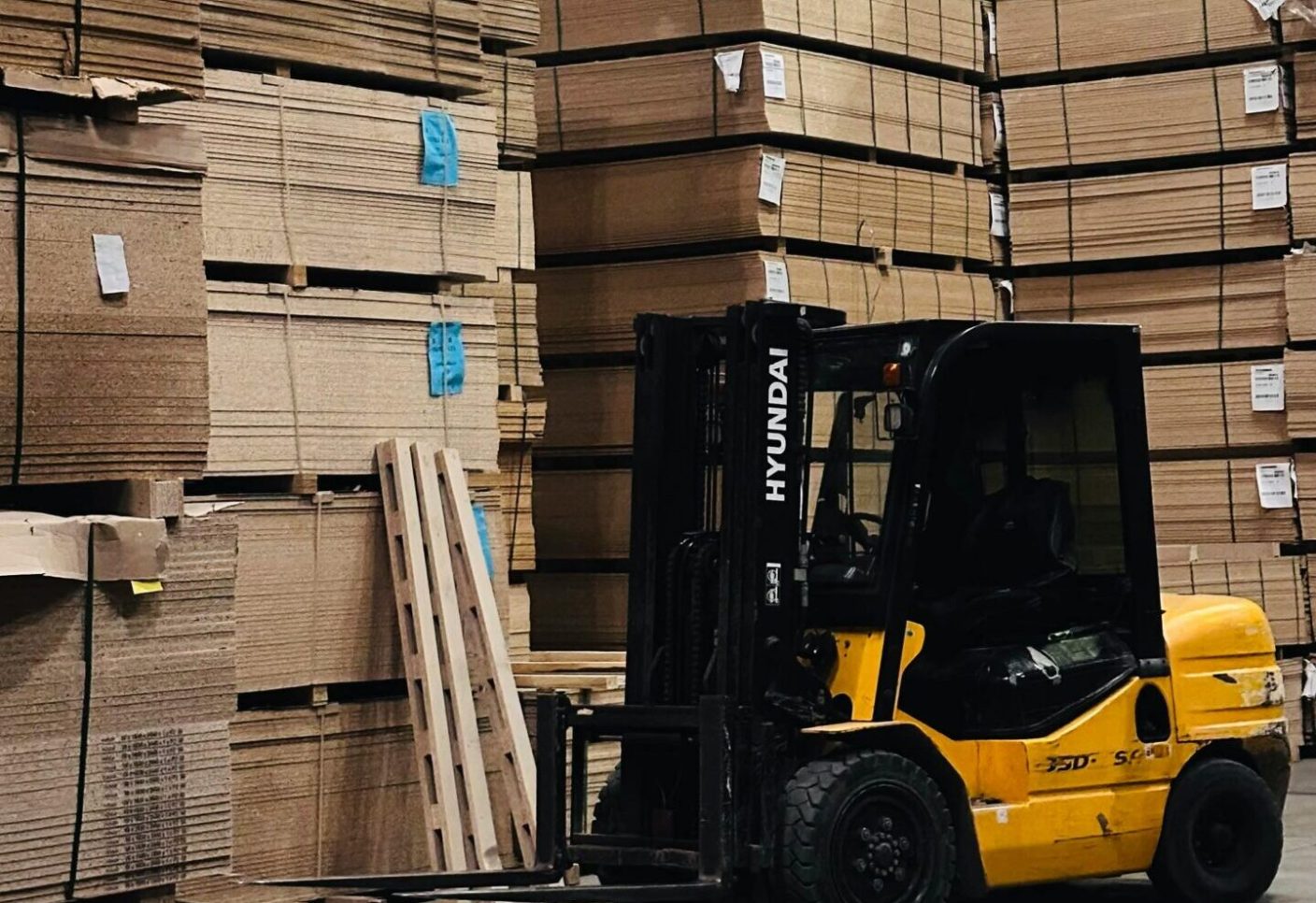 A yellow forklift organizes large stacks of cardboard boxes in a spacious warehouse.